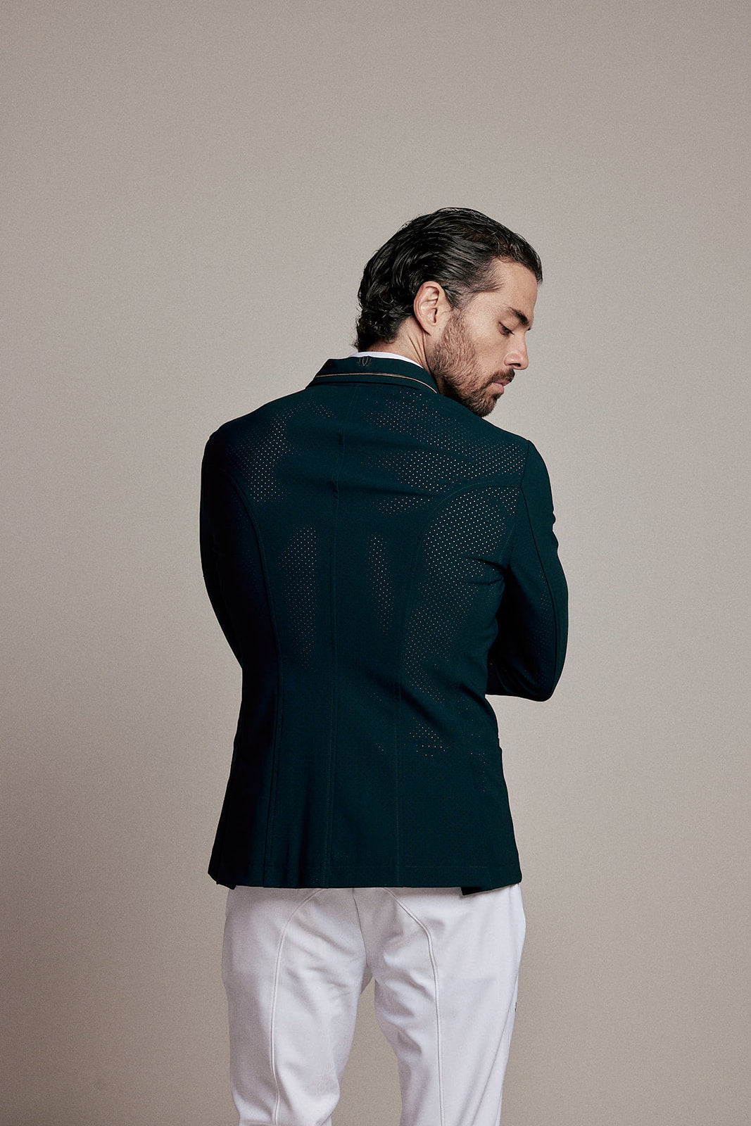 A man shown from the back wearing a dark teal textured blazer and white trousers, standing against a solid beige studio background.