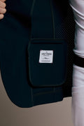 Close-up of a dark navy equestrian vest with a front pouch pocket featuring a white 'VEL+REIS' brand label, worn by a model in white riding pants.