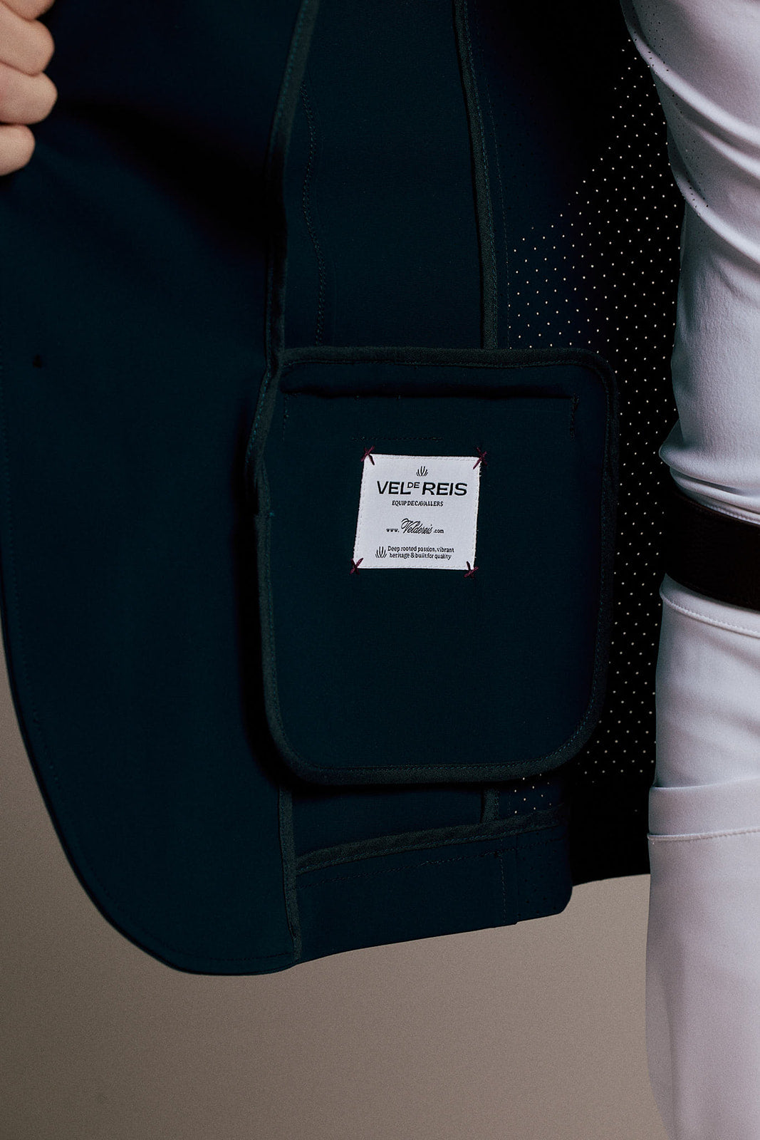 Close-up of a dark navy equestrian vest with a front pouch pocket featuring a white 'VEL+REIS' brand label, worn by a model in white riding pants.