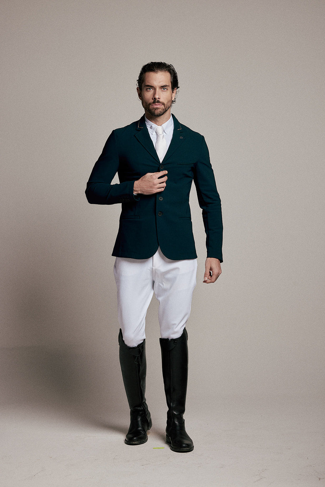 A man modeling a dark teal equestrian blazer, white riding breeches, a white dress shirt, and black riding boots against a neutral beige studio background.