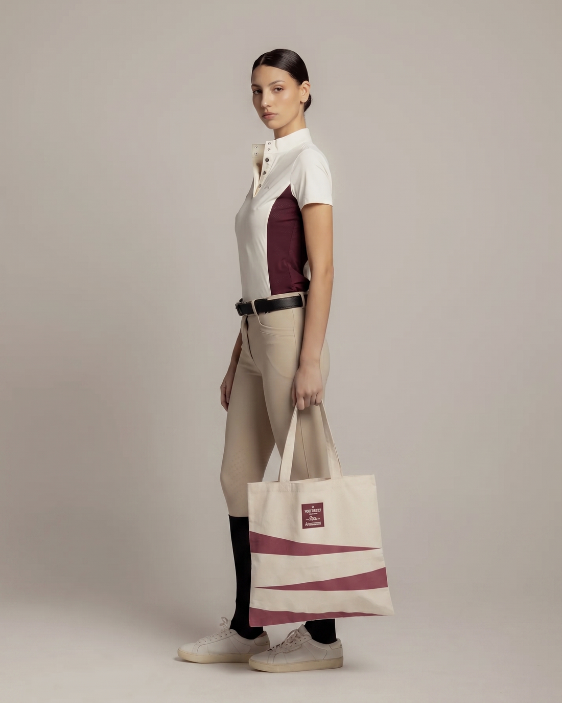 A model stands in profile wearing a white and burgundy short-sleeve polo shirt, beige riding breeches, black socks, and white sneakers, while holding a cream canvas tote bag with burgundy diagonal stripes and a small red label.