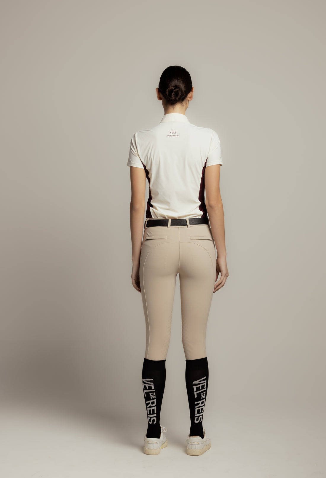 A woman modeling a short-sleeve polo shirt in off-white with a small embroidered logo on the chest, paired with beige riding pants and a black belt, against a solid light gray background.