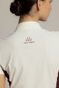 Close-up back view of a woman wearing a white collared golf shirt with a burgundy Velt-Reis logo printed on the upper back, set against a neutral light gray background.