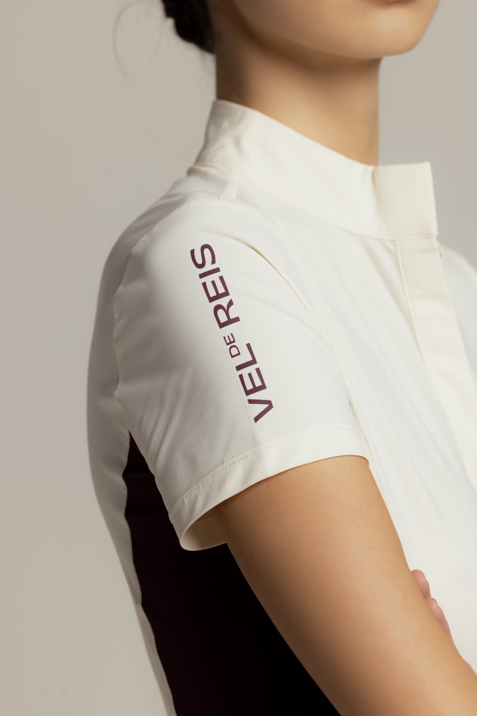 Close-up of a model wearing a white short-sleeve collared shirt with 'VEL D'ESPRIT' printed vertically in dark purple on the upper sleeve, set against a neutral light gray background.