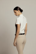 A woman shown from the back wearing a white short-sleeve polo shirt with dark side panels and a small logo on the upper back, paired with beige riding pants and a black belt, against a plain light gray studio background.