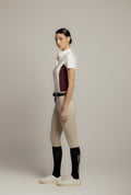 A woman stands in profile wearing a white short-sleeve polo shirt with a dark burgundy side panel, beige riding breeches, a black belt, black knee-high socks, and white sneakers against a plain light gray studio background.