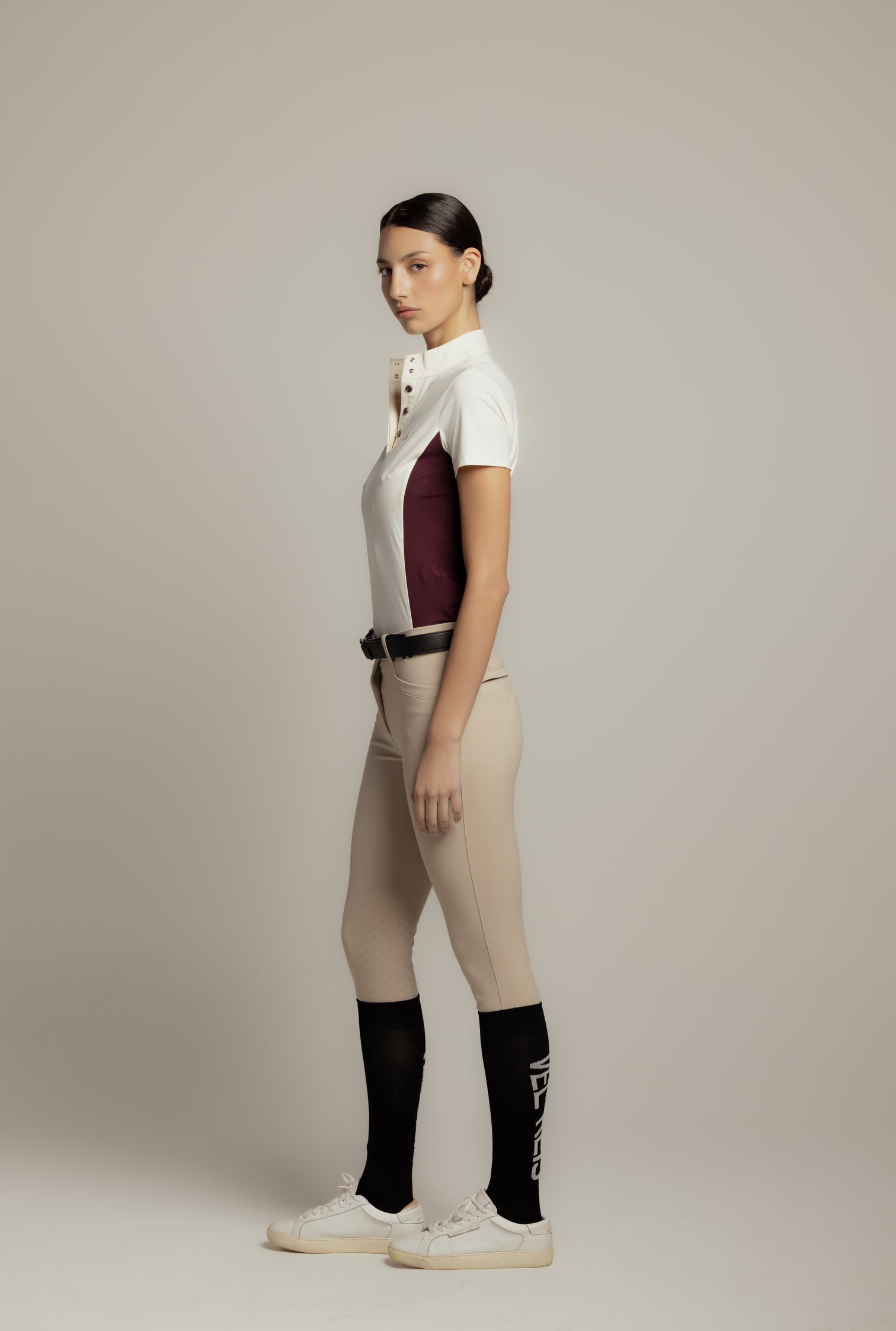 A woman stands in profile wearing a white short-sleeve polo shirt with a dark burgundy side panel, beige riding breeches, a black belt, black knee-high socks, and white sneakers against a plain light gray studio background.