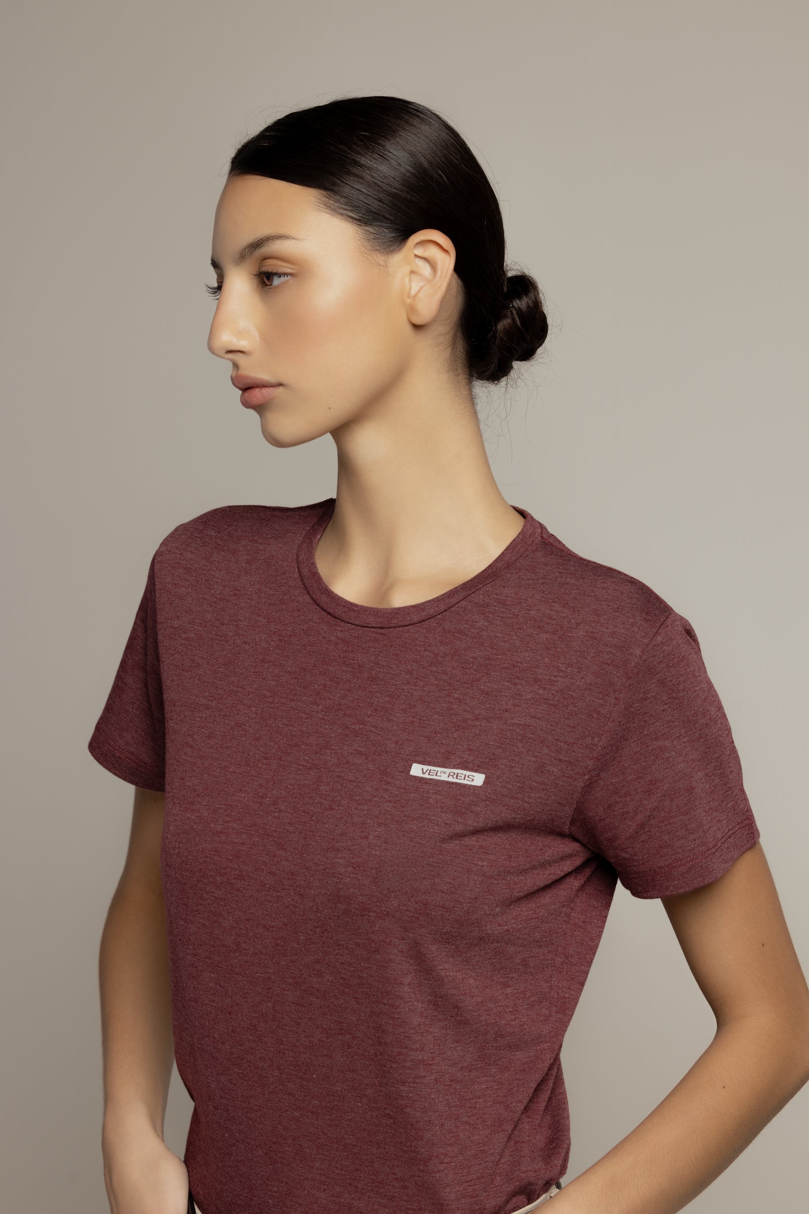A woman wearing a short-sleeve crewneck t-shirt in heathered burgundy, shown from the waist up against a solid light gray background.