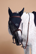 A close-up of a white and gray spotted horse wearing a dark navy fly veil and brown leather bridle against a solid beige background.