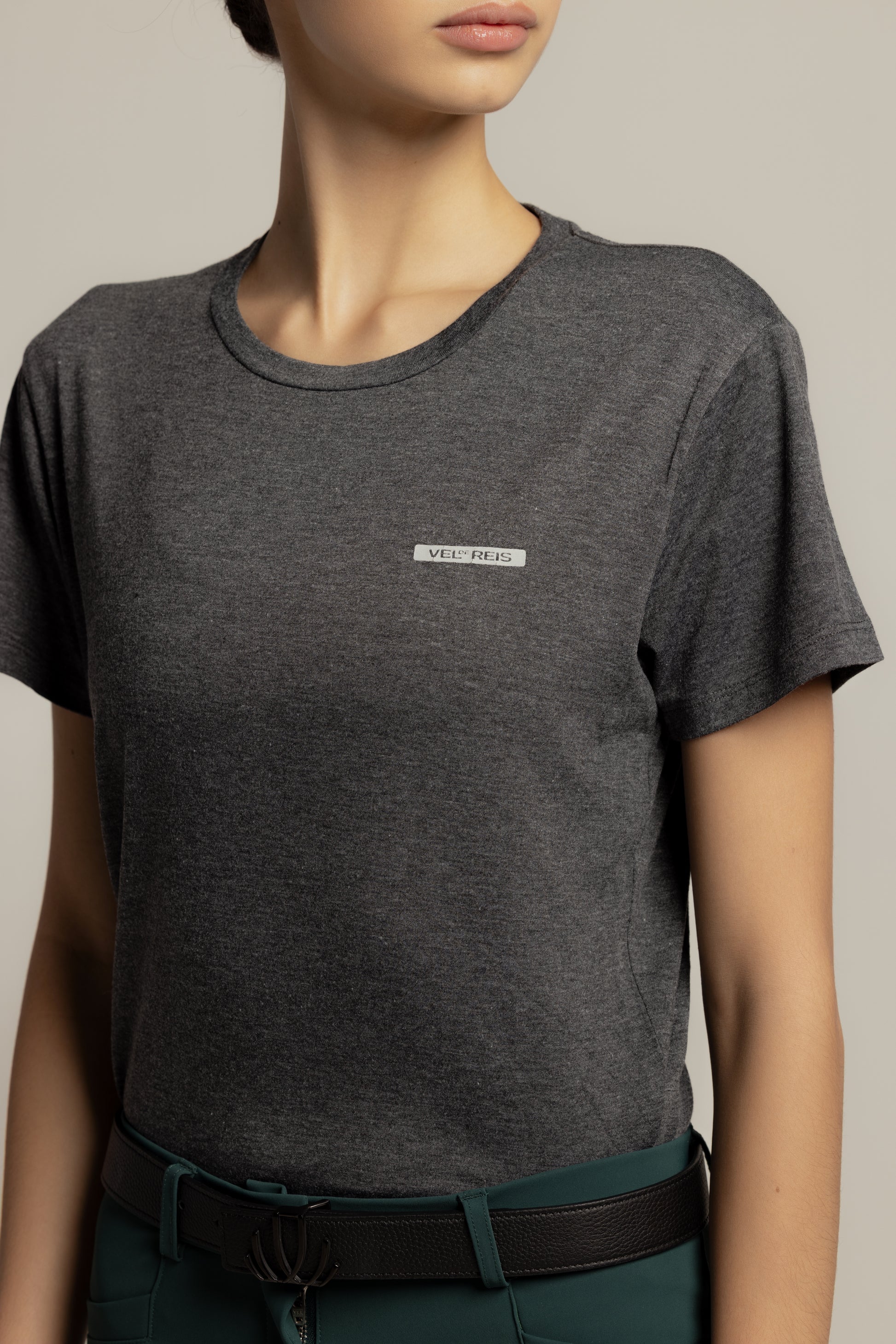 A close-up of a model wearing a dark heather gray crewneck t-shirt with a small white rectangular logo patch on the left chest, paired with dark green trousers and a black belt.