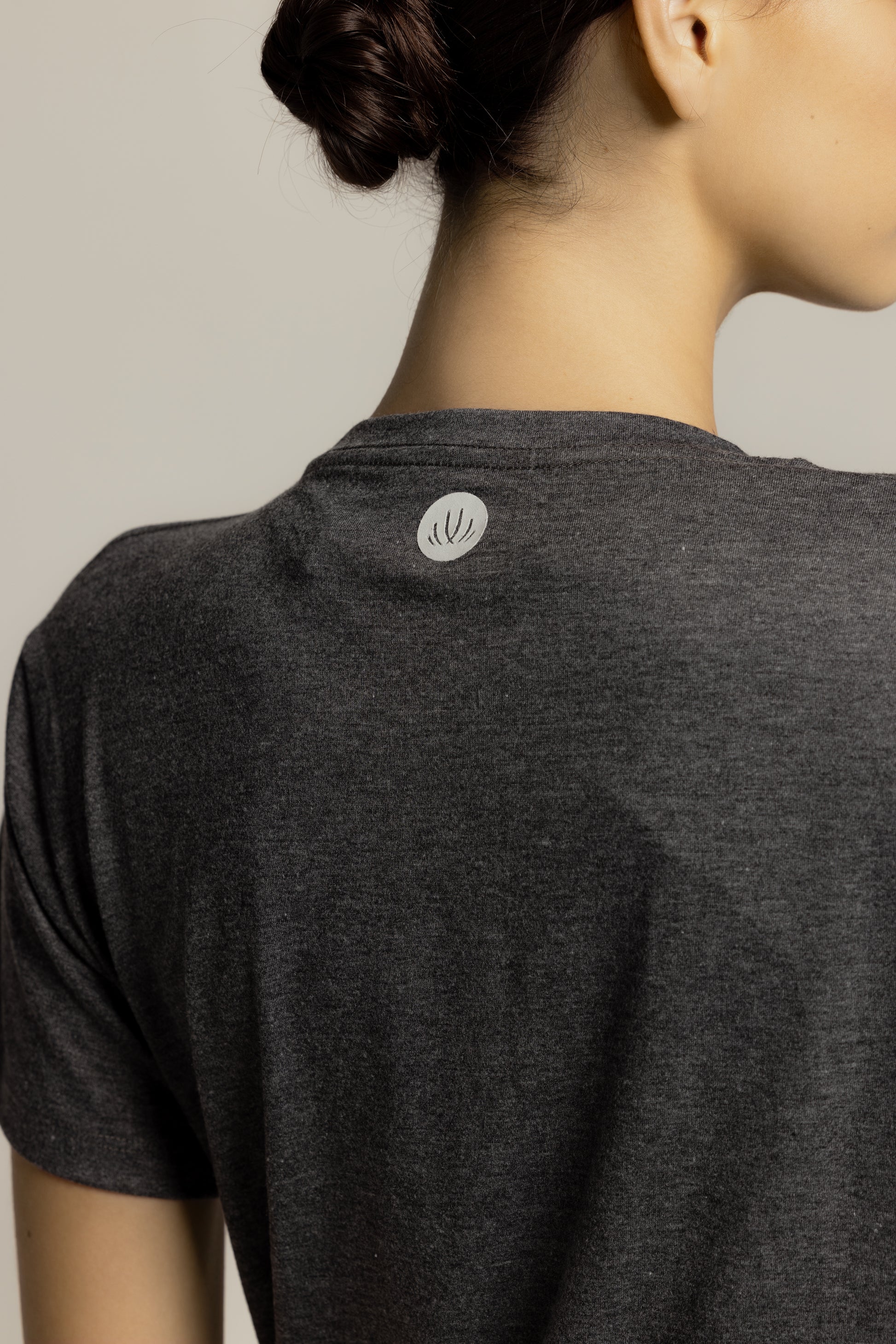 Close-up back view of a person wearing a dark heather gray short-sleeve T-shirt, showing a small circular white logo centered above the neckline.