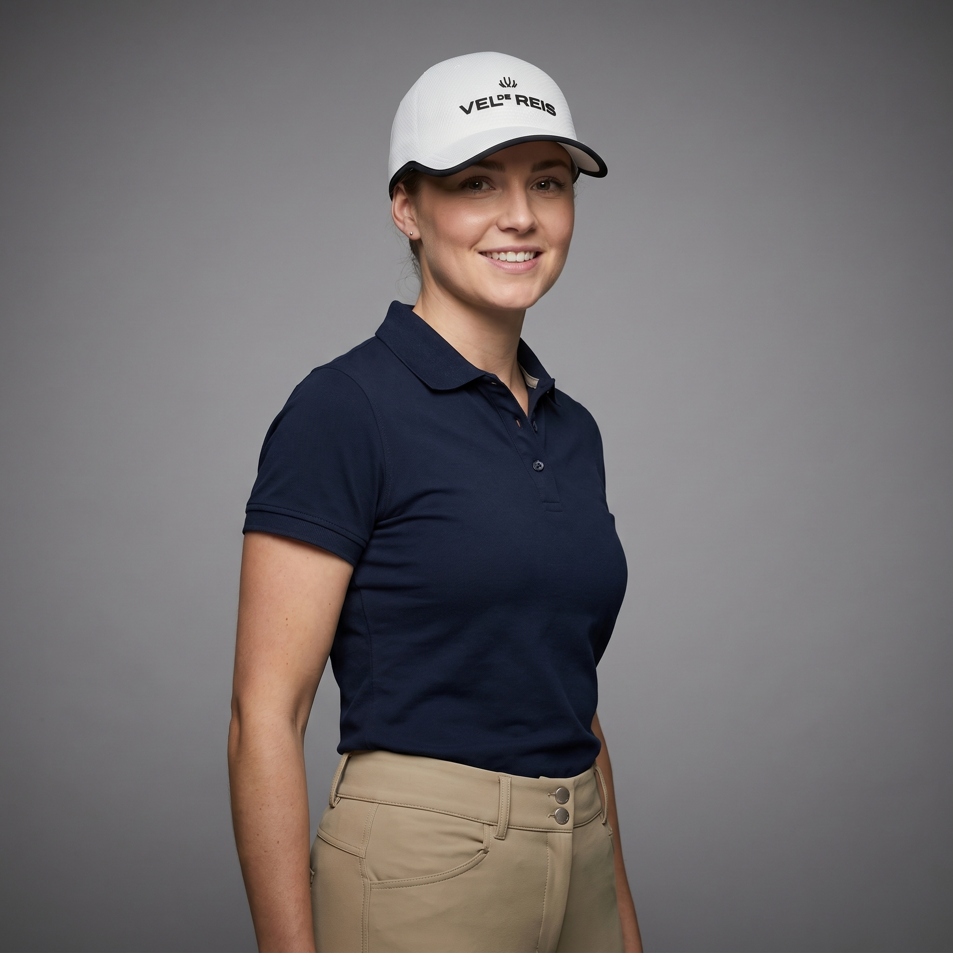 Woman wearing a navy polo shirt and beige pants with a white cap on a gray background