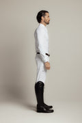 A man in profile wearing a white long-sleeve shirt, white riding breeches, and black riding boots, standing against a plain light gray studio background.
