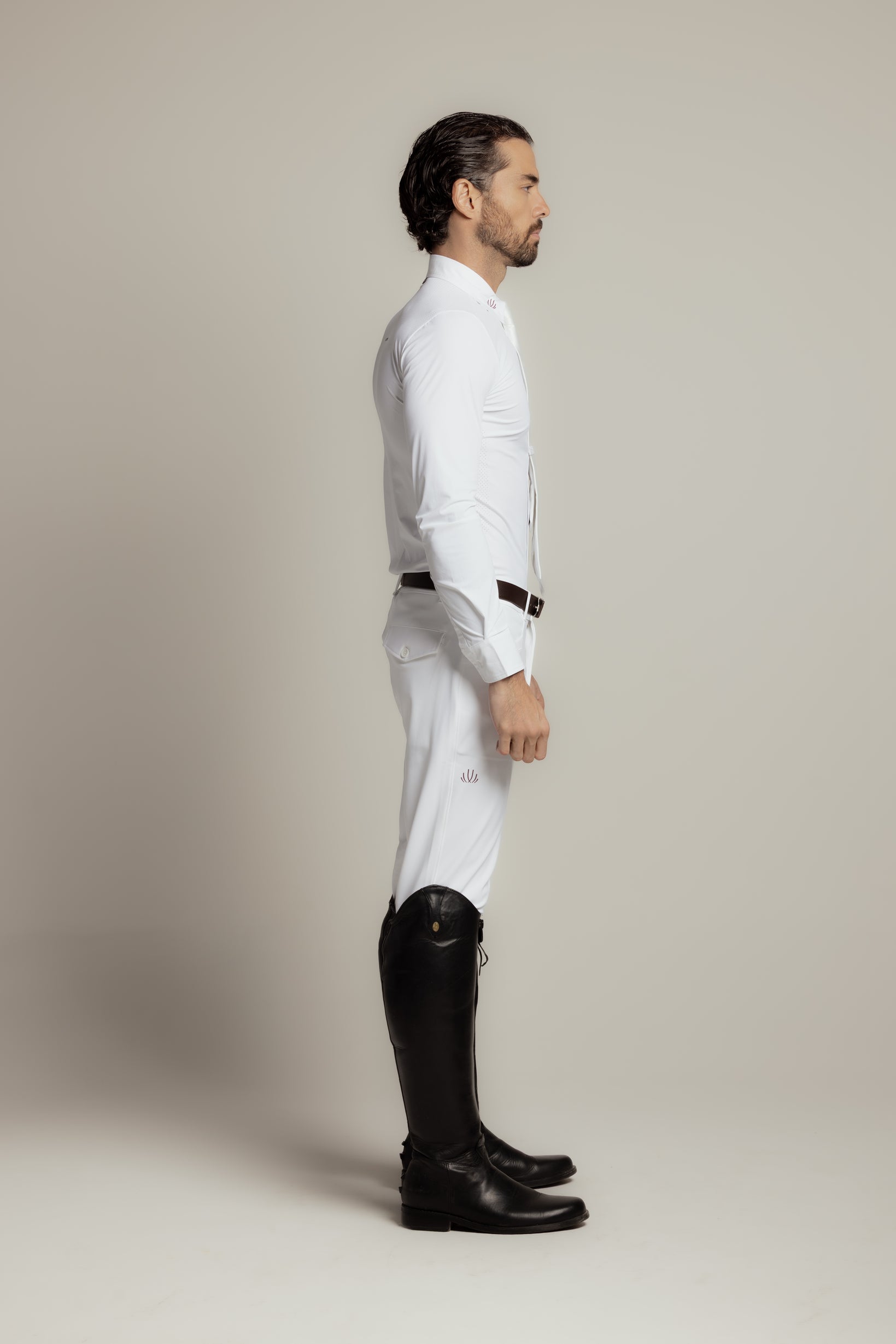 A man in profile wearing a white long-sleeve shirt, white riding breeches, and black riding boots, standing against a plain light gray studio background.