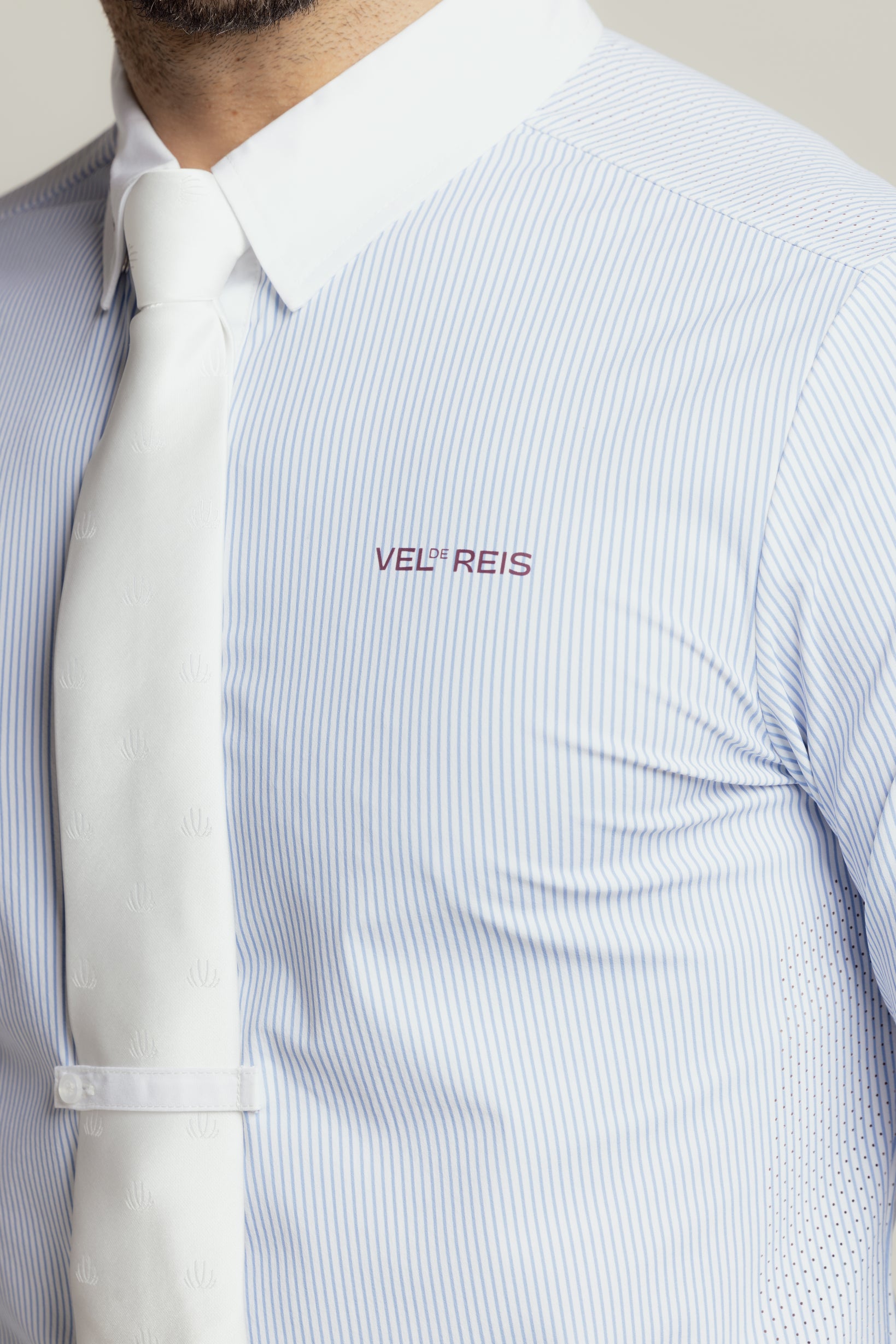 Close-up of a man wearing a light blue and white pinstriped dress shirt with a white tie. The shirt features the brand name 'VEL REIS' embroidered in burgundy on the chest.