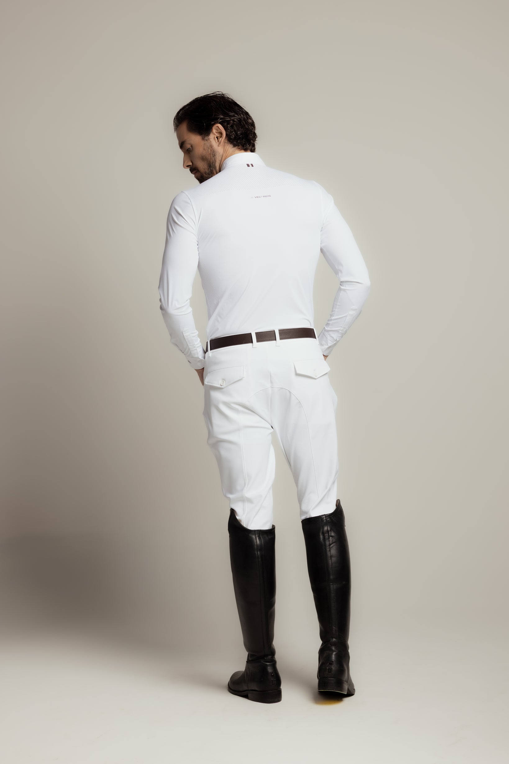 A man shown from the back wearing a white long-sleeve equestrian shirt, white riding breeches, a dark brown belt, and black riding boots against a neutral studio background.