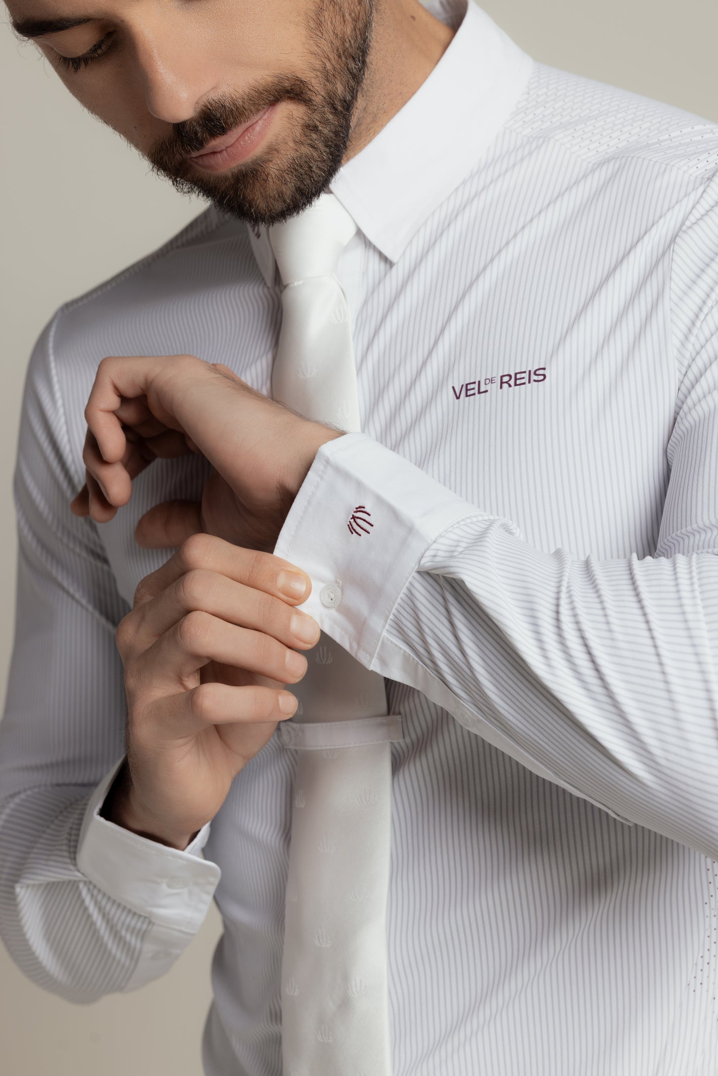 A close-up of a man wearing a white long-sleeve dress shirt with subtle vertical pinstripes, a white tie, and adjusting the cuff. The shirt features the brand name 'VEL REIS' embroidered on the chest.