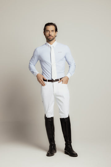 A man modeling a light blue long-sleeve dress shirt, white tie, white riding breeches, a dark brown leather belt, and black riding boots against a neutral light gray studio background.