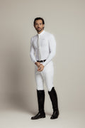 A man modeling a white long-sleeve dress shirt, white tie, white riding breeches, and black riding boots against a neutral beige studio background.