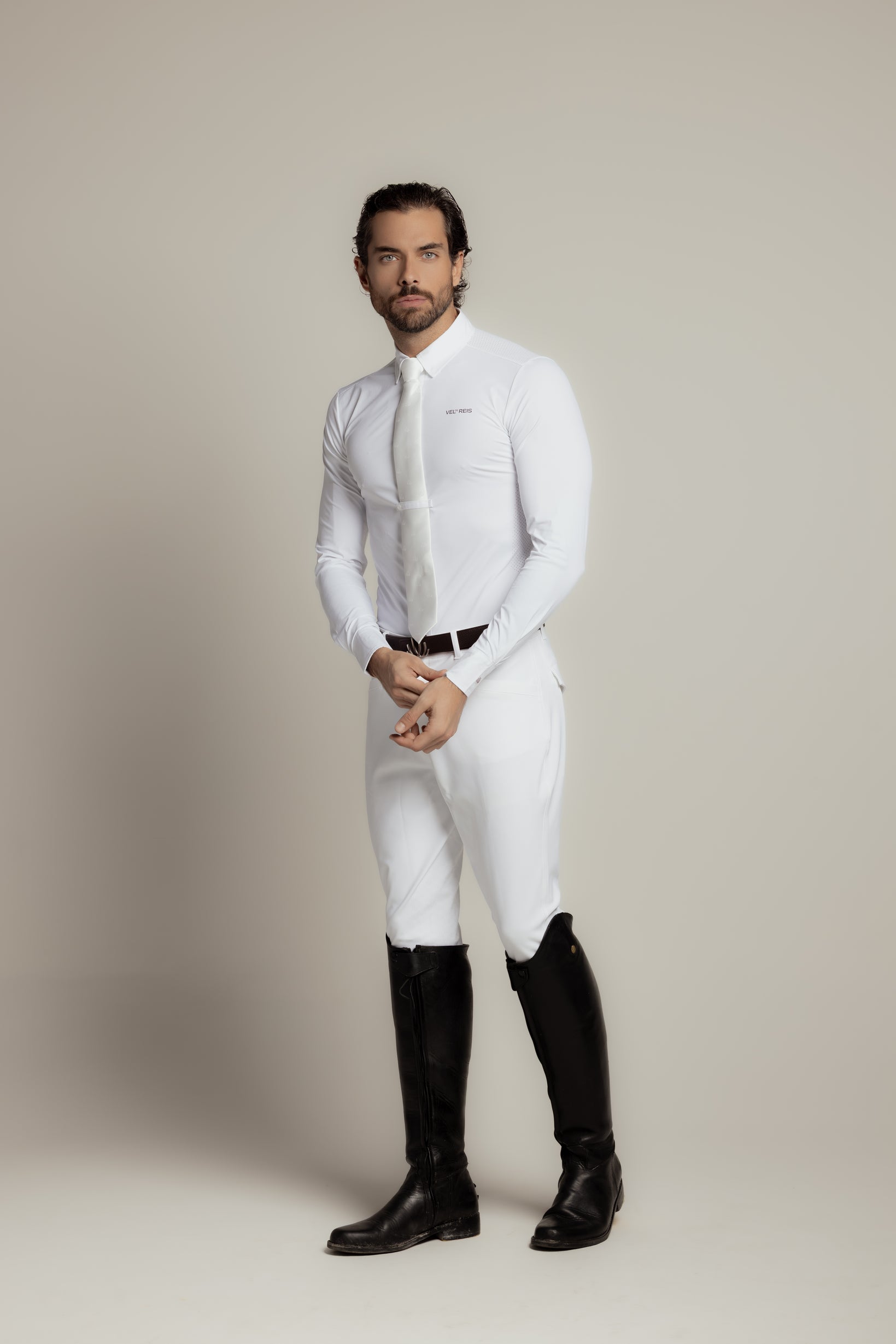 A man modeling a white long-sleeve dress shirt, white tie, white riding breeches, and black riding boots against a neutral beige studio background.