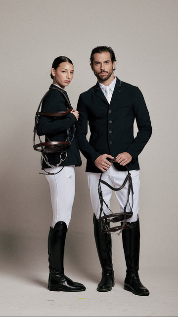 A man and a woman in equestrian attire pose against a neutral beige studio background. Both wear dark navy jackets, white breeches, and tall black riding boots, with leather bridles hanging from their shoulders.