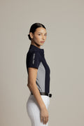 A woman modeling a short-sleeve athletic jersey in navy and light gray, paired with white riding pants, against a solid light gray studio background. The jersey features the brand name 'Velo Rides' printed vertically on the sleeve.