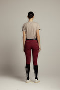 A model shown from the back wearing high-waisted burgundy riding tights with black socks featuring white branding, a light gray t-shirt, and white sneakers, set against a plain light gray studio background.