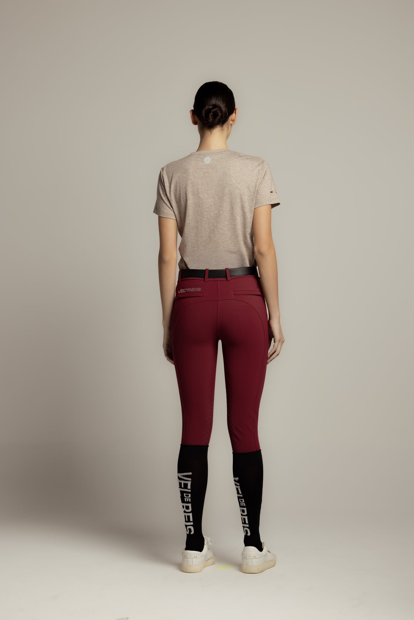 A model shown from the back wearing high-waisted burgundy riding tights with black socks featuring white branding, a light gray t-shirt, and white sneakers, set against a plain light gray studio background.