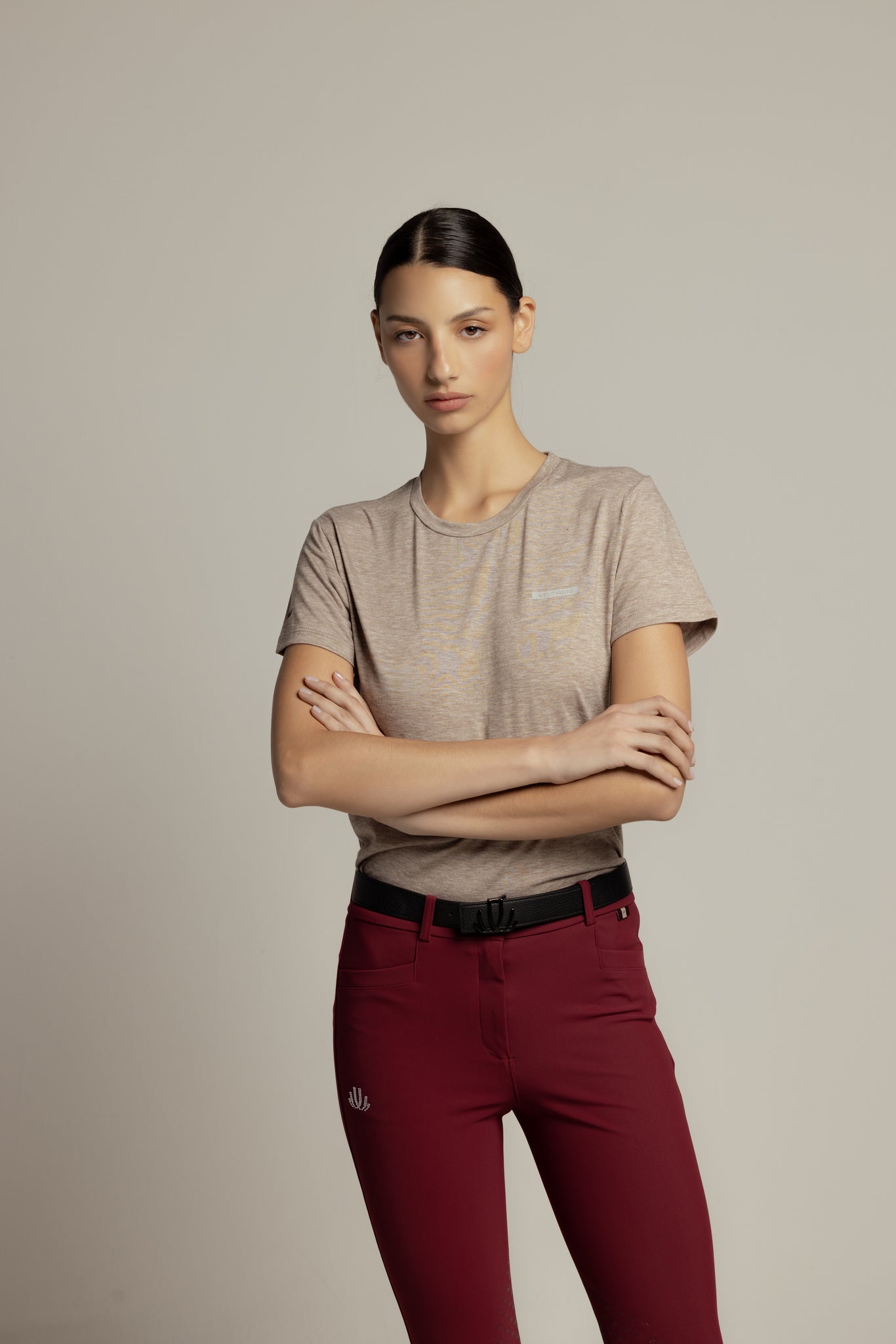 A woman modeling a light beige short-sleeve t-shirt and dark red slim-fit pants with a black belt, standing against a solid light gray background.