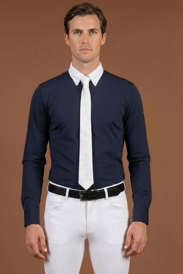 Man wearing a navy long-sleeve shirt with a white Pre-tied show Tie from VEL DE REIS, white competition breeches, and a black belt against a brown background.