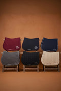 Six horse saddle pads in different colors and textures arranged in two rows on small wooden stands against a warm brown background. The top row features curved saddle pads in burgundy, navy blue, and cream, while the bottom row features rectangular saddle pads in gray and black.