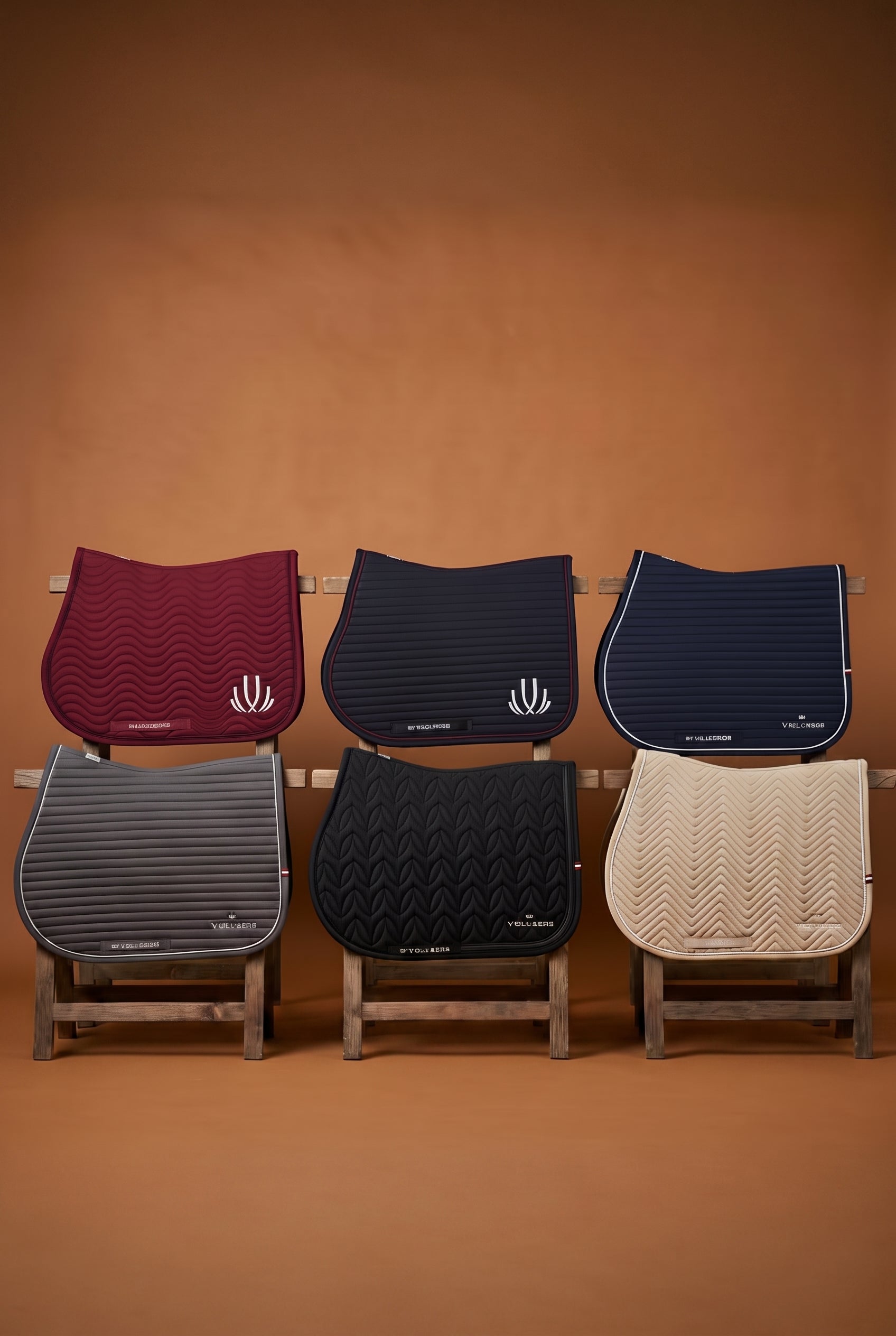 Six horse saddle pads in different colors and textures arranged in two rows on small wooden stands against a warm brown background. The top row features curved saddle pads in burgundy, navy blue, and cream, while the bottom row features rectangular saddle pads in gray and black.