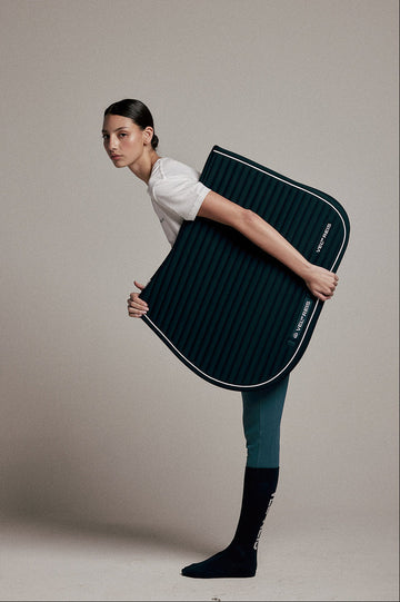 A woman in a white t-shirt, teal leggings, and black socks holds a dark teal ribbed horse saddle pad with white piping and a small white logo, posed against a solid beige studio background.
