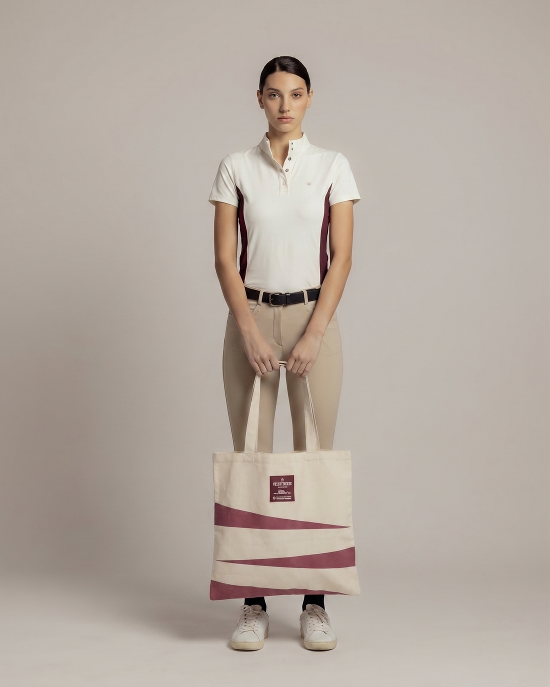A model stands in profile wearing a white and burgundy short-sleeve polo shirt, beige riding breeches, black socks, and white sneakers, while holding a cream canvas tote bag with burgundy diagonal stripes and a small red label.