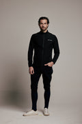 A man modeling a black zip-up athletic jacket, black track pants, and white sneakers against a neutral beige studio background.