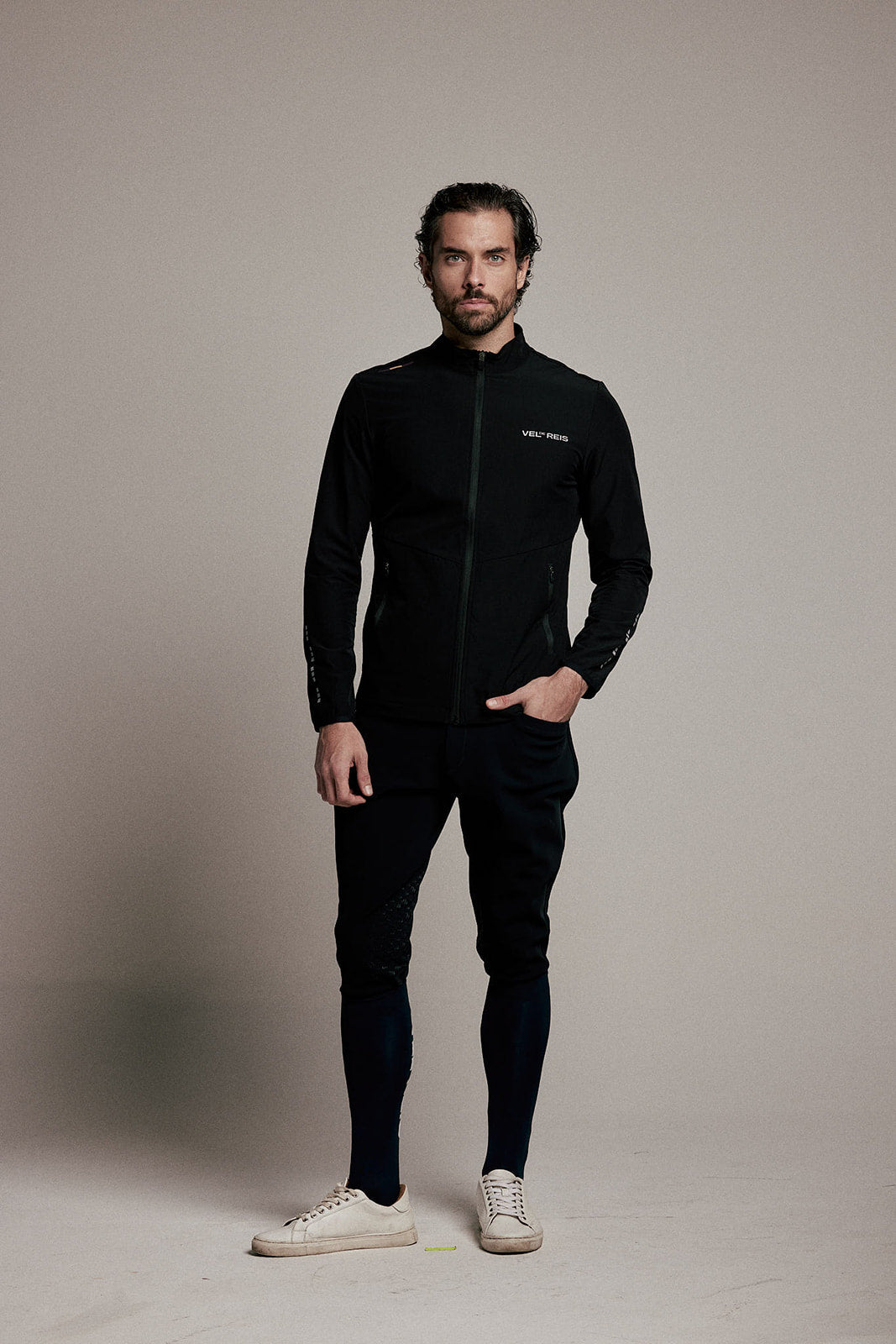 A man modeling a black zip-up athletic jacket, black track pants, and white sneakers against a neutral beige studio background.