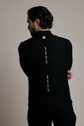 A man viewed from behind wearing a black long-sleeve athletic top with a vertical dotted white line graphic down the back and a small circular logo near the collar, against a plain light beige studio background.
