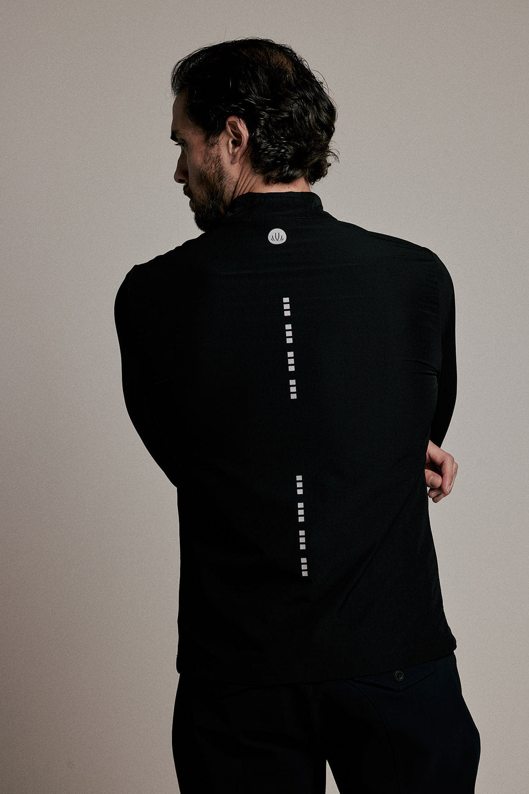 A man viewed from behind wearing a black long-sleeve athletic top with a vertical dotted white line graphic down the back and a small circular logo near the collar, against a plain light beige studio background.