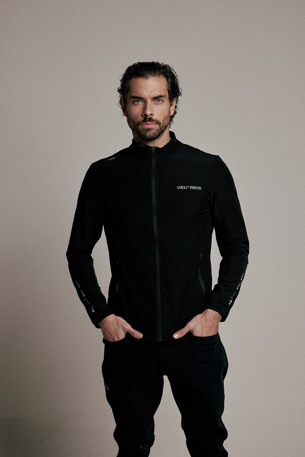 A man wearing a black zip-up athletic jacket with a white 'VENI REIS' logo on the chest, paired with black pants, standing against a plain beige studio background.