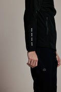 A close-up side view of a person wearing a black athletic jacket and matching black pants against a plain beige studio background. The jacket has a vertical row of reflective square-like accents down one sleeve and a small white logo on the pants.
