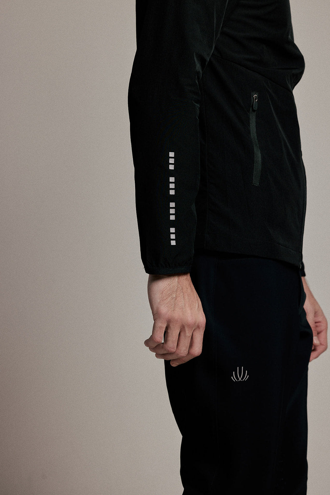 A close-up side view of a person wearing a black athletic jacket and matching black pants against a plain beige studio background. The jacket has a vertical row of reflective square-like accents down one sleeve and a small white logo on the pants.