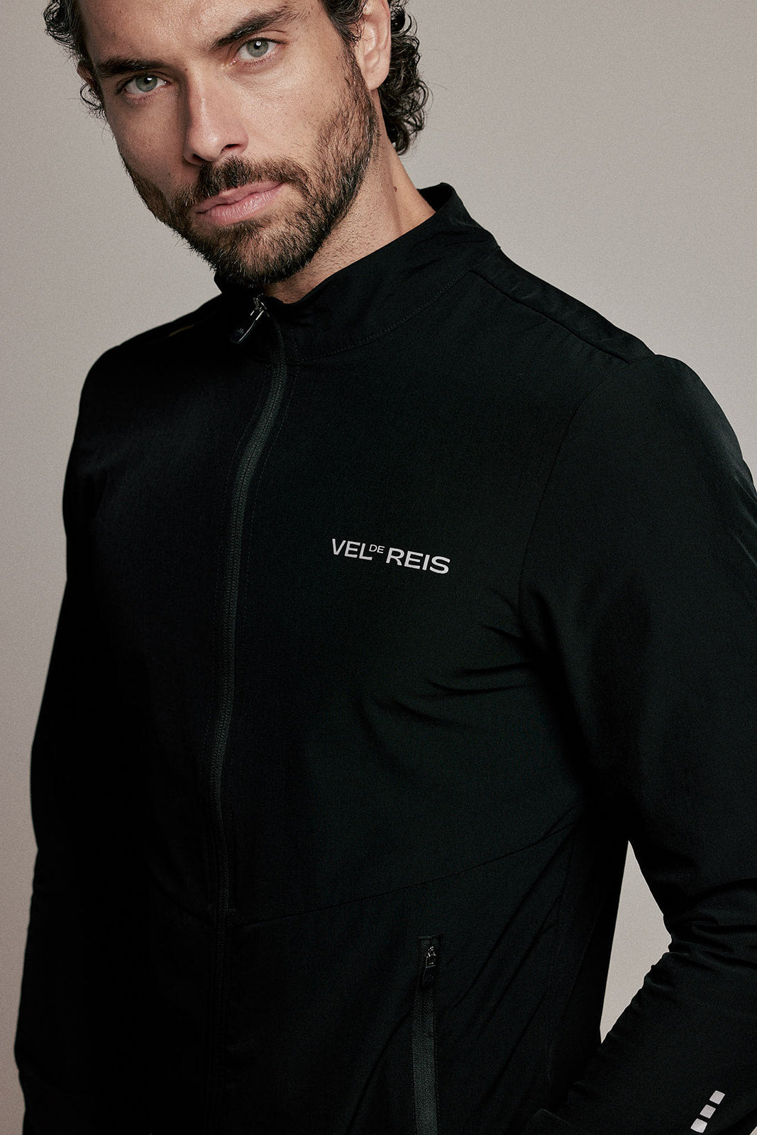 A close-up of a man wearing a black athletic zip-up jacket with the brand name 'VEL'REIS' printed in white on the left chest, set against a plain light gray background.