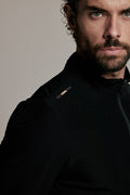 A close-up portrait of a man wearing a black athletic zip-up jacket against a plain light gray studio background.