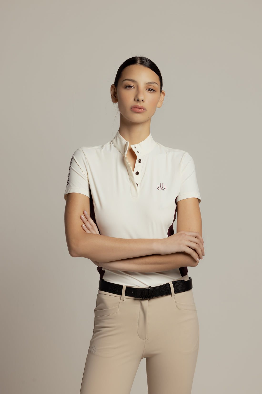 A woman modeling a short-sleeve polo shirt in off-white with a small embroidered logo on the chest, paired with beige riding pants and a black belt, against a solid light gray background.