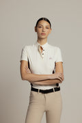 A woman modeling a short-sleeve polo shirt in off-white with a small embroidered logo on the chest, paired with beige riding pants and a black belt, against a solid light gray background.