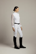 A woman stands in profile wearing a fitted white long-sleeve mock-neck riding shirt with sheer sleeves, white riding breeches, a black belt, and black riding boots against a plain warm gray studio background.