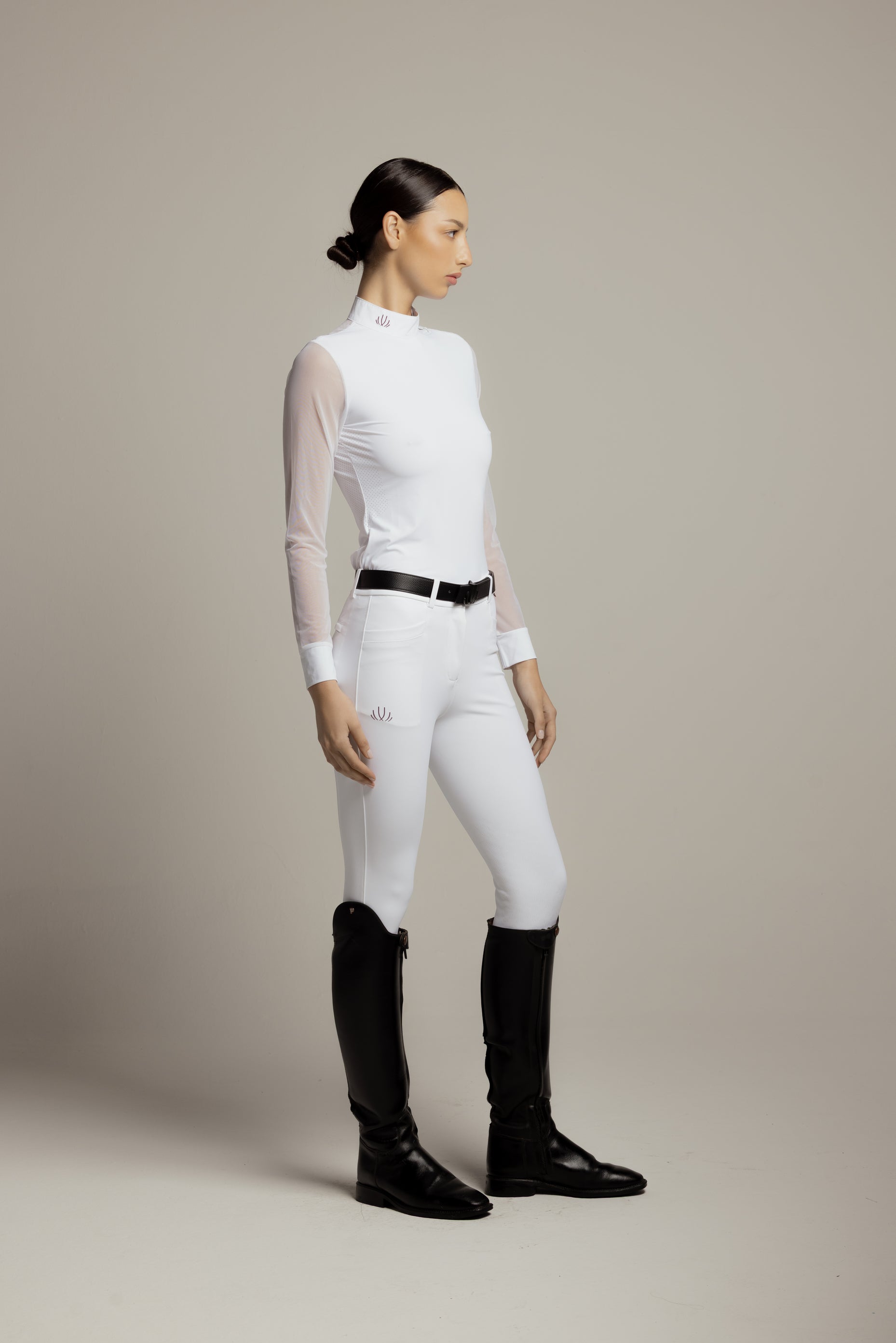 A woman stands in profile wearing a fitted white long-sleeve mock-neck riding shirt with sheer sleeves, white riding breeches, a black belt, and black riding boots against a plain warm gray studio background.