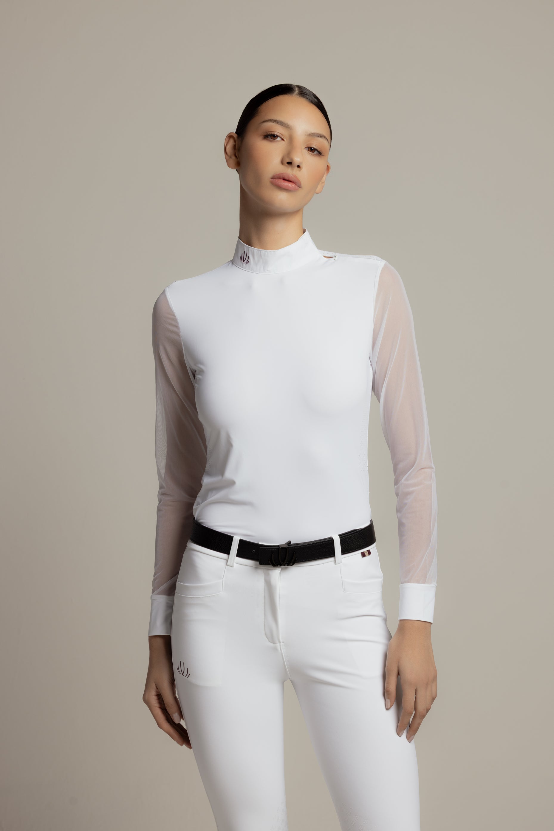 A model wearing a white mock-neck riding shirt with sheer long sleeves, paired with white riding pants and a black belt, against a neutral beige studio background.