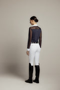 A woman shown from the back wearing a navy sheer-paneled long-sleeve top, white riding breeches, a black belt, and black riding boots against a plain beige studio background.