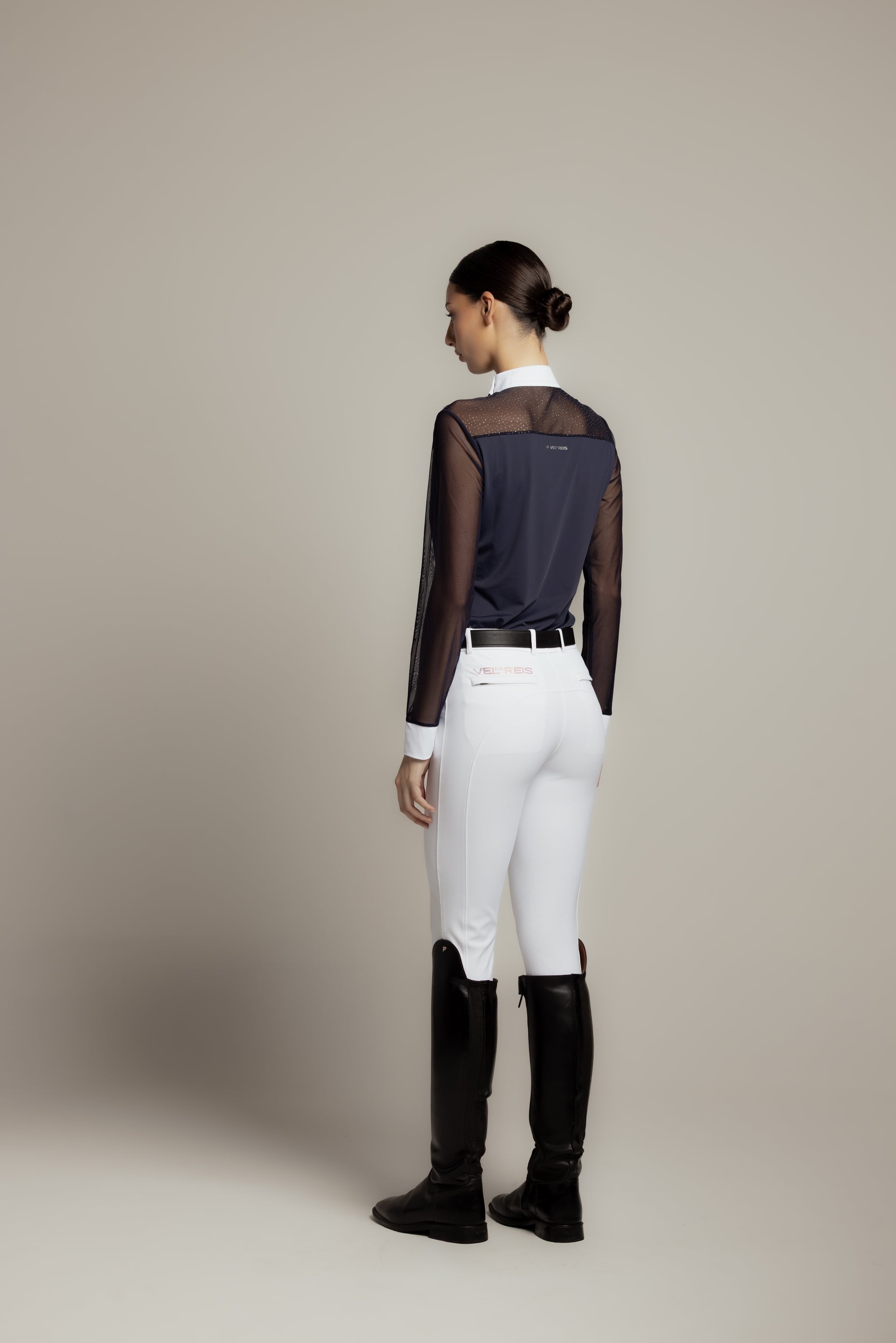 A woman shown from the back wearing a navy sheer-paneled long-sleeve top, white riding breeches, a black belt, and black riding boots against a plain beige studio background.
